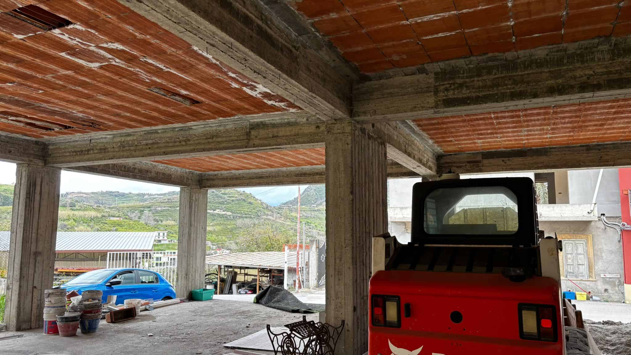 Cantiere edile con struttura in cemento e mattoni, vista su un paesaggio collinare, con veicoli e materiali da costruzione, pronto per la fase di finitura e ristrutturazione.