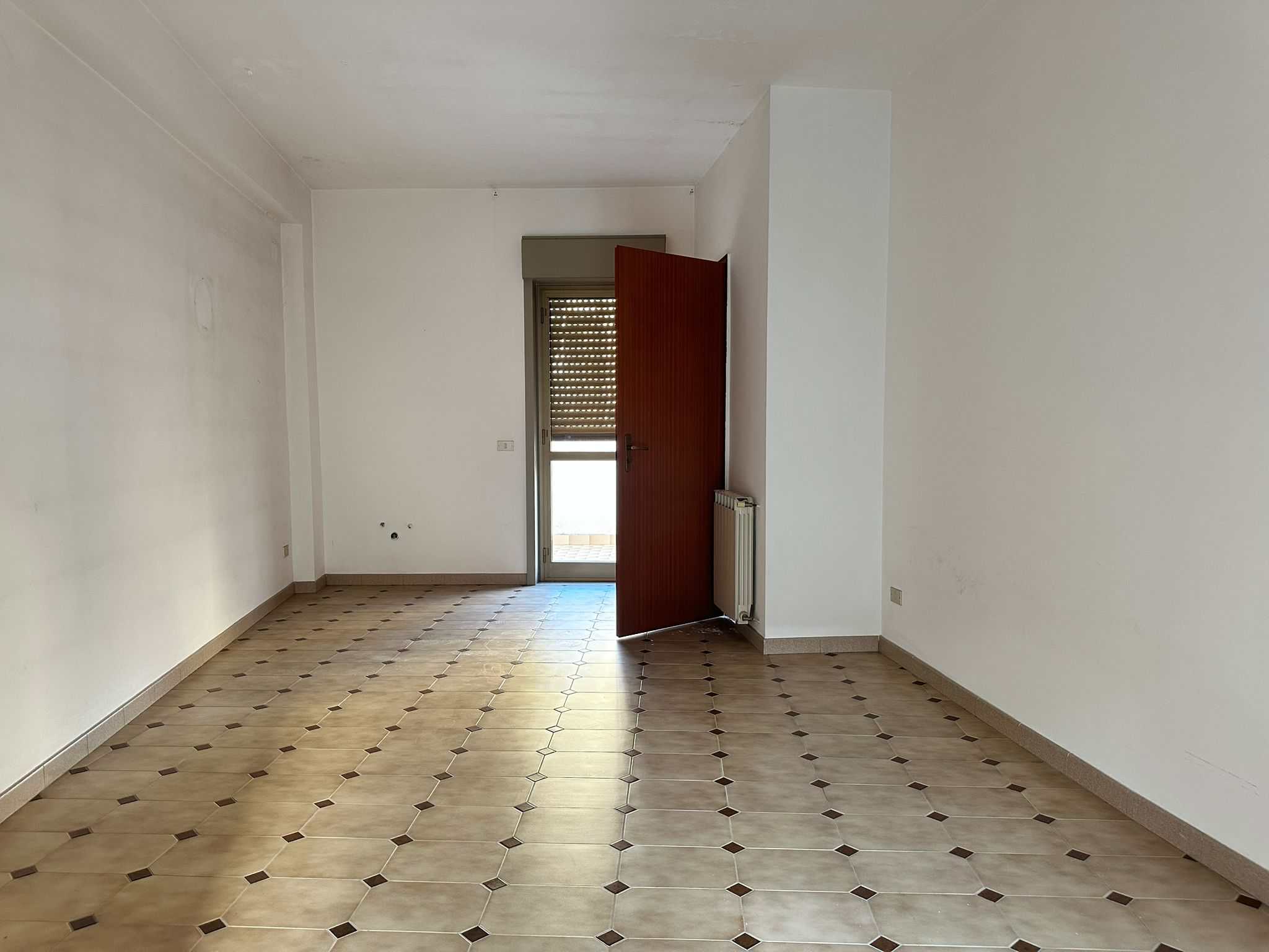 Appartamento vuoto con pareti bianche e pavimento in piastrelle, vista sulla porta d'ingresso e balcone, ideale per ristrutturazione e arredamento.