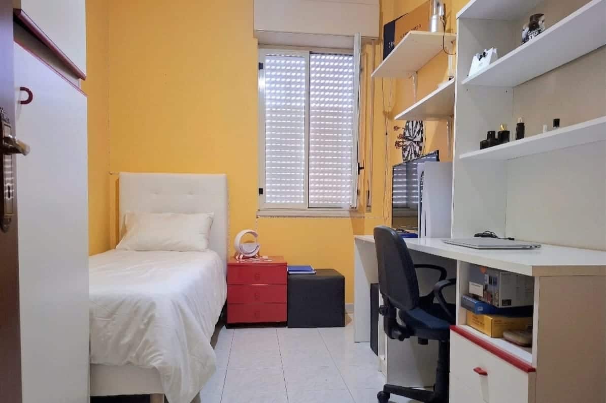 Una stanza singola luminosa con pareti gialle, letto singolo con biancheria bianca, scrivania con sedia ergonomica e scaffali con oggetti vari, ideale per studenti o professionisti.