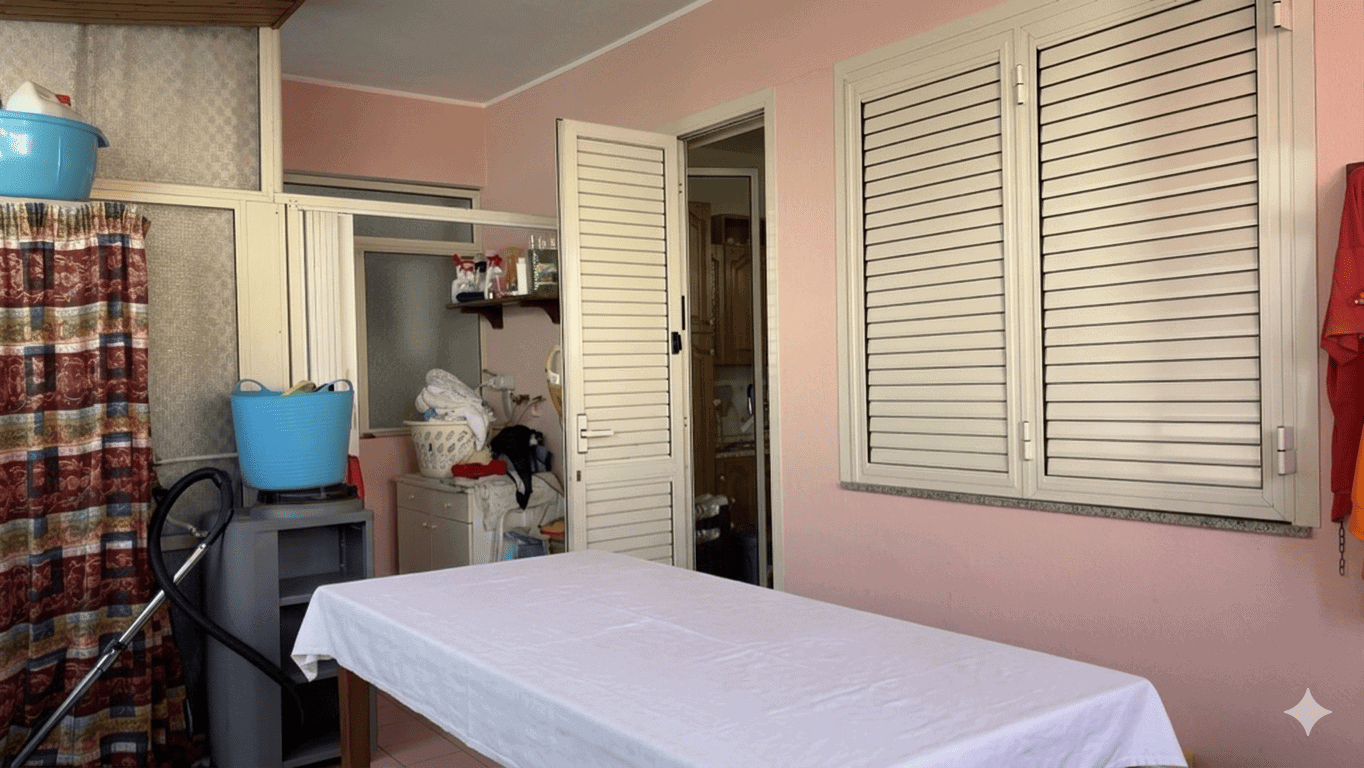 Tavolo da stiro in una stanza con pareti rosa, finestra con persiane e armadio a battente, ambiente domestico ordinato e funzionale.