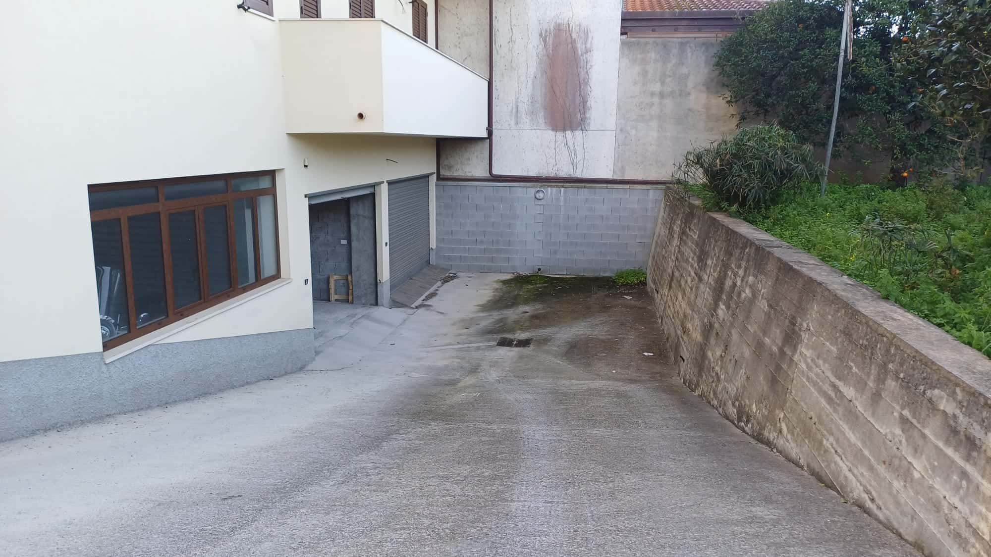 Ampio cortile esterno con superficie in cemento e accesso garage, situato accanto a un edificio residenziale moderno con finestre grandi e pareti bianche.