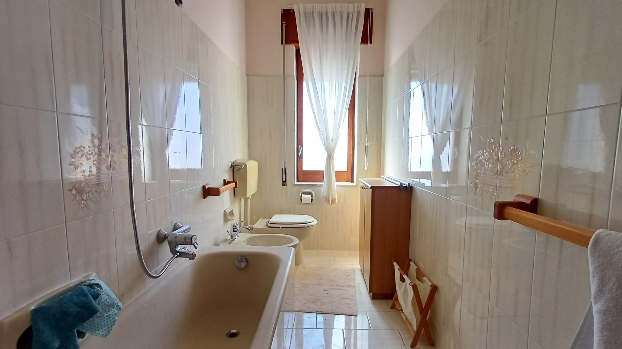Lavabo e vasca da bagno in un bagno luminoso con finestra, tende bianche e arredi in legno, ambiente pulito e accogliente, ideale per il comfort quotidiano.