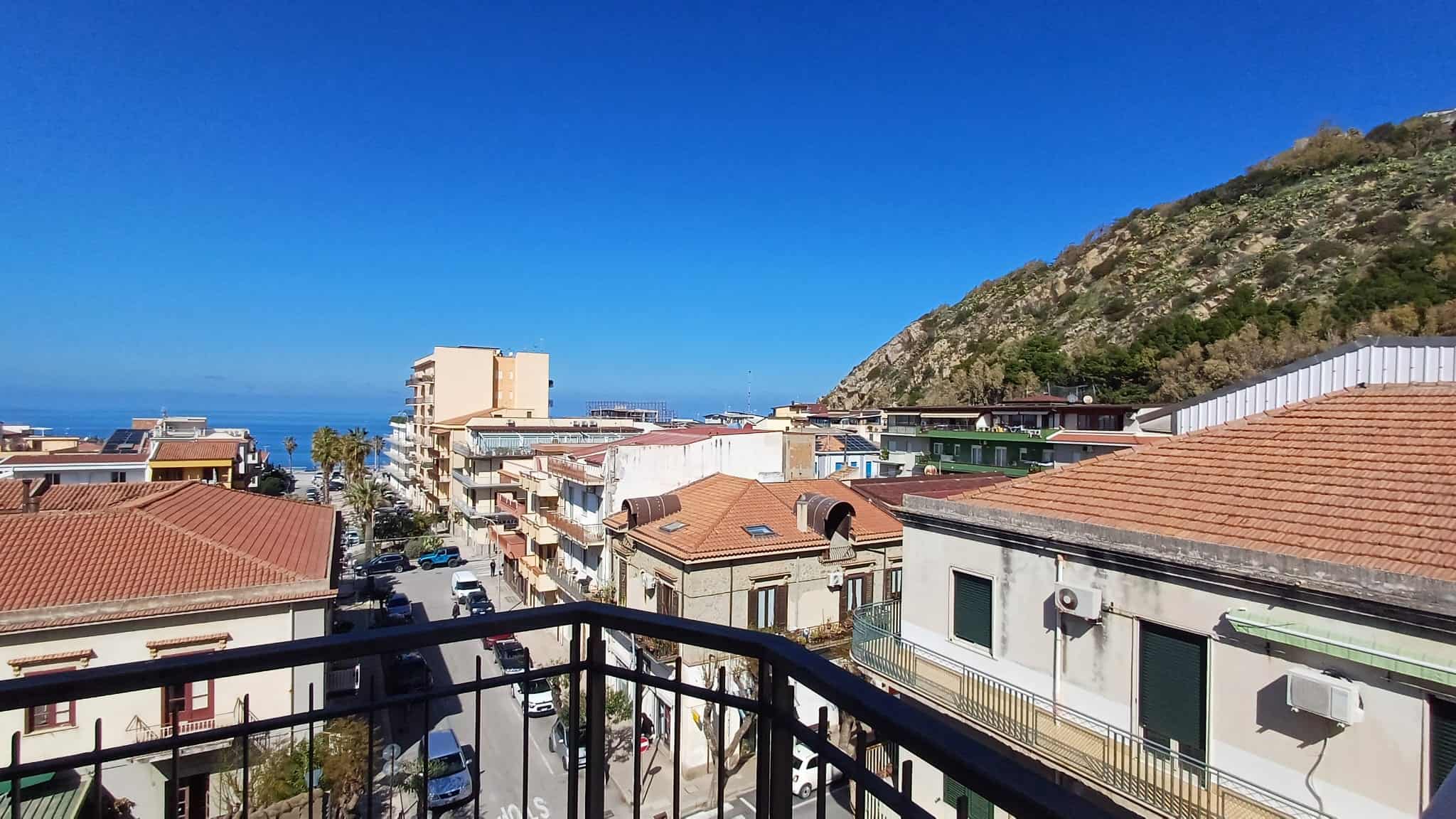 Balcone con vista sul mare e sulle case di un quartiere residenziale vicino alla costa, con un monte sullo sfondo, cielo sereno e edifici con tetti in terracotta.