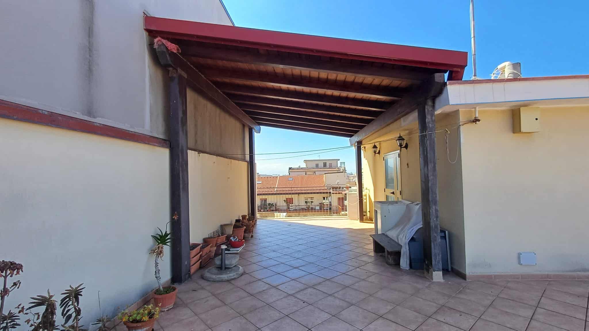 Il tettoia in legno e metallo su un terrazzo urbano con piante in vaso, vista sulla città, sotto un cielo azzurro.