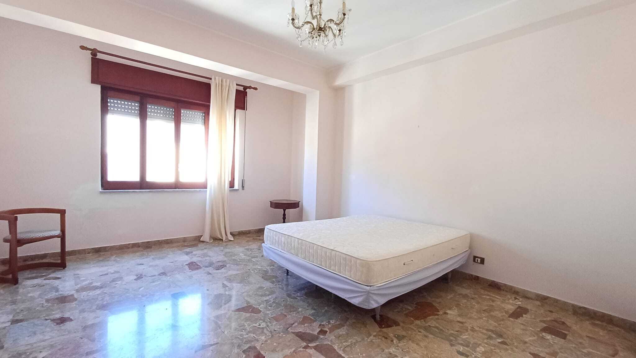 Letto matrimoniale in una stanza luminosa con pareti bianche, finestra con tende e pavimento in marmo, ambiente semplice e spazioso.