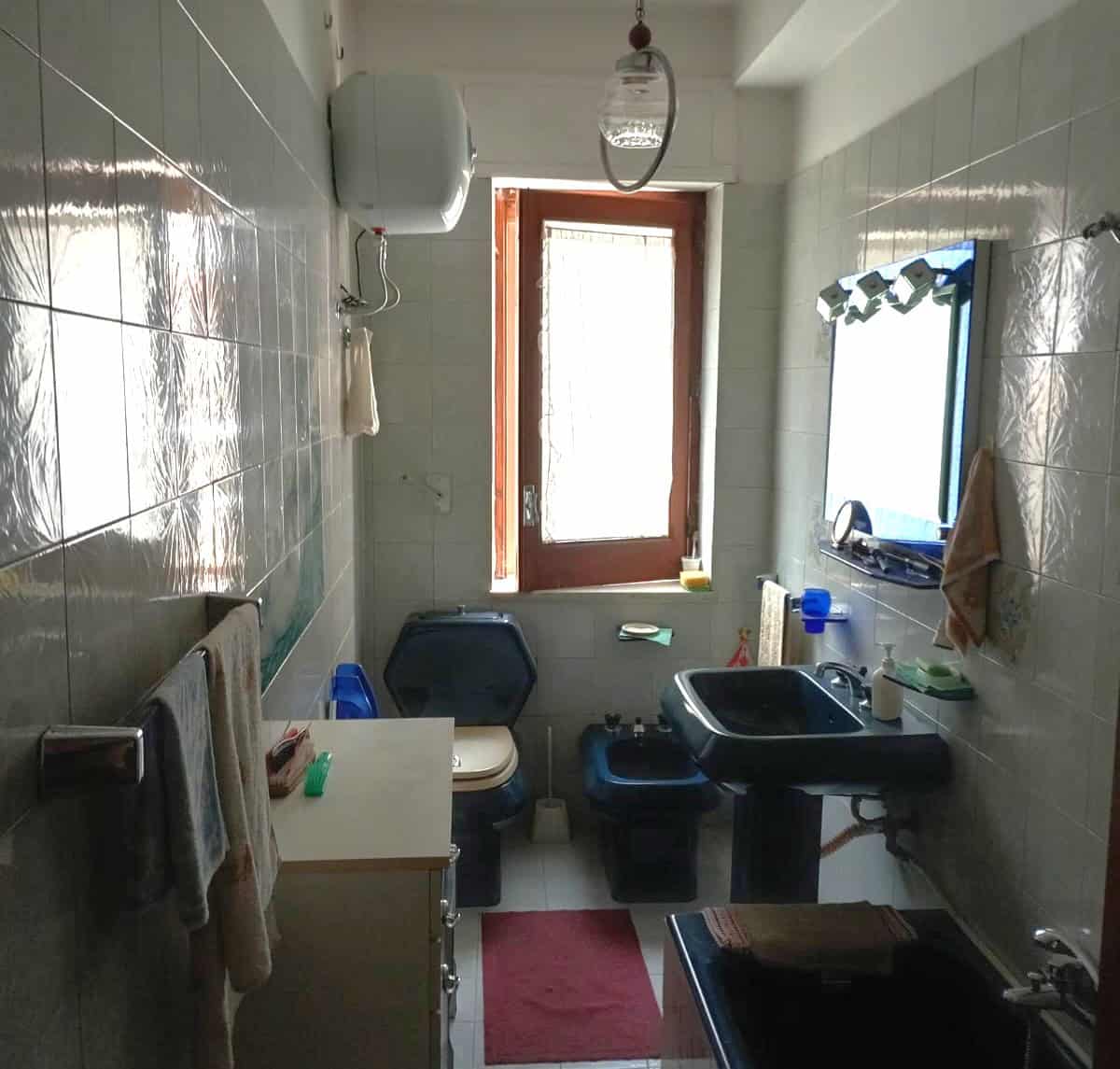 Lavabo e servizi igienici in un bagno di piccole dimensioni con finestra, piastrelle chiare, specchio e accessori essenziali, ideale per soluzioni di arredo funzionali e pratiche.