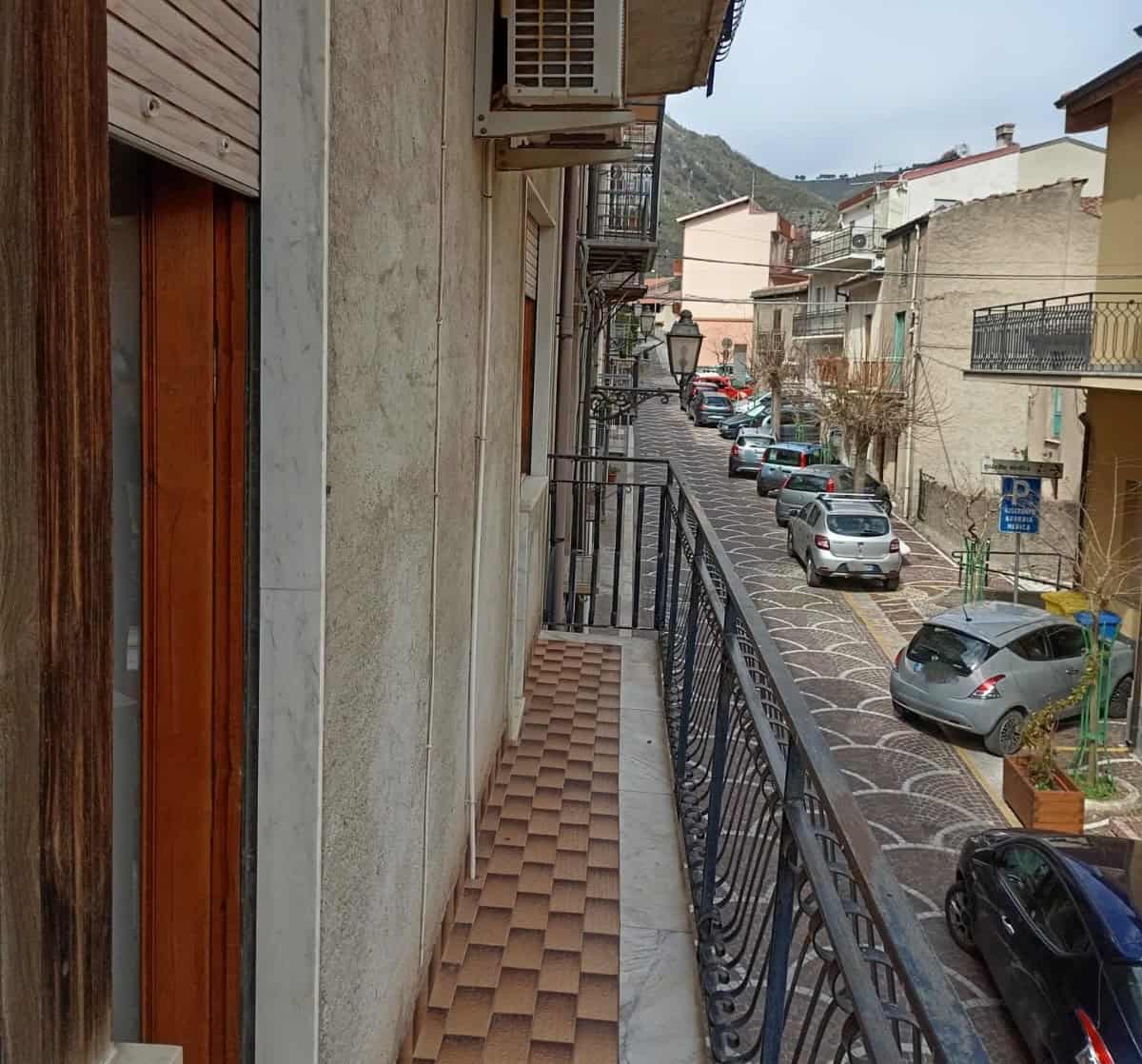 Balcone di un edificio in un quartiere residenziale con vista sulla strada e sulle auto parcheggiate, in un contesto urbano con edifici colorati e colline sullo sfondo.