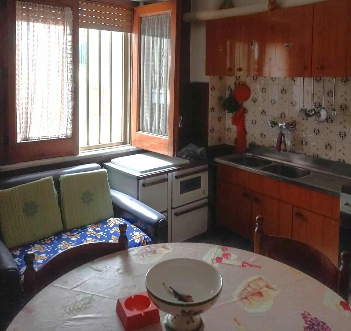 Cucina abitabile con finestra, mobili in legno, tavolo rotondo e divano con cuscini verdi, ambiente accogliente e luminoso.