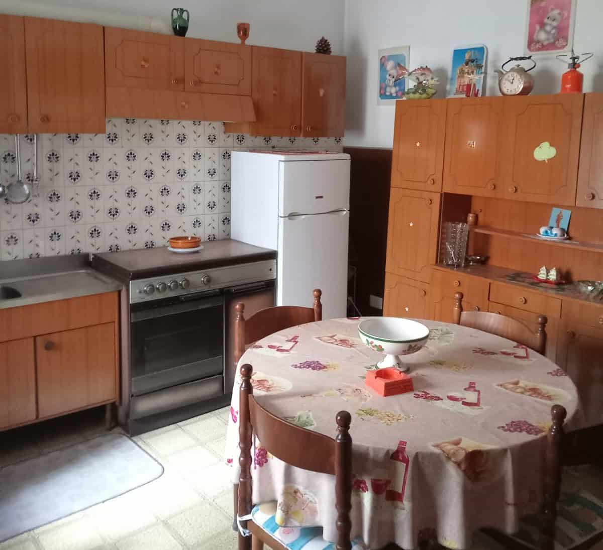Pannello di cucina vintage con mobili in legno, frigorifero bianco e tavolo rotondo con tovaglia decorata, ambiente domestico accogliente e tradizionale.