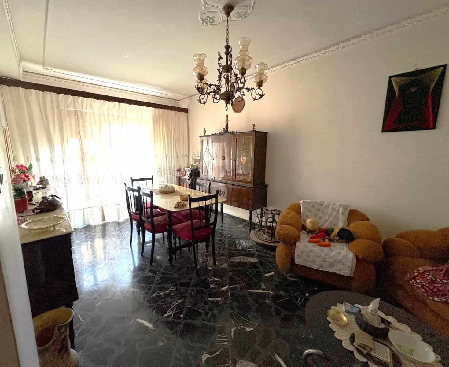 Interno soggiorno con mobili classici, tavolo da pranzo, divani e lampadario in stile vintage, illuminato da luce naturale.