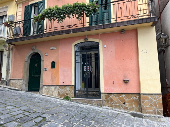 Portone d'ingresso in ferro battuto con porta pedonale, facciata colorata in rosa e giallo, con balcone e finestre con persiane verdi, situata in una strada acciottolata.