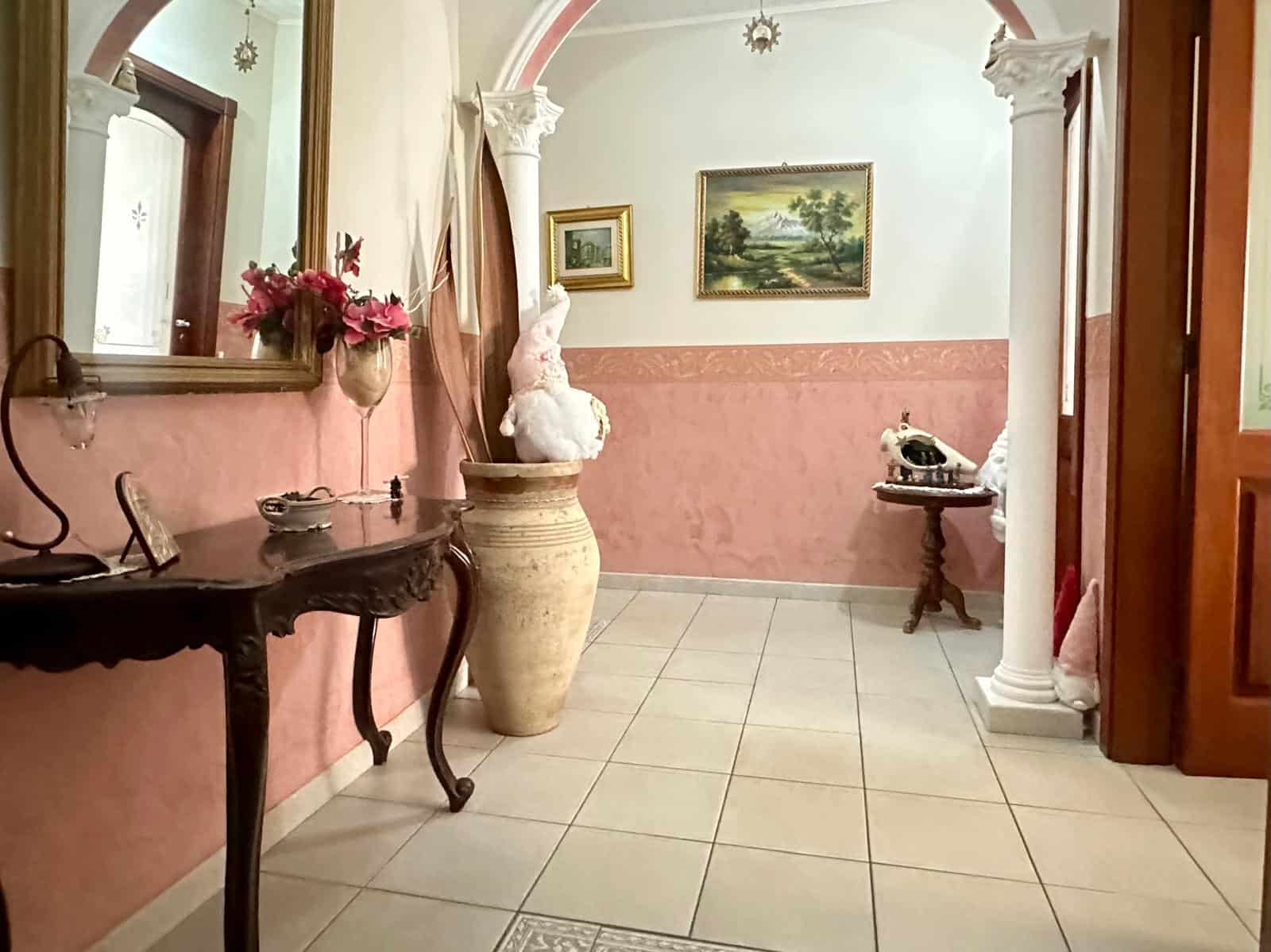 Elegante ingresso con pareti rosa, decorato con fiori e opere d'arte, arricchito da colonne bianche e mobili classici, creando un'atmosfera accogliente e raffinata.