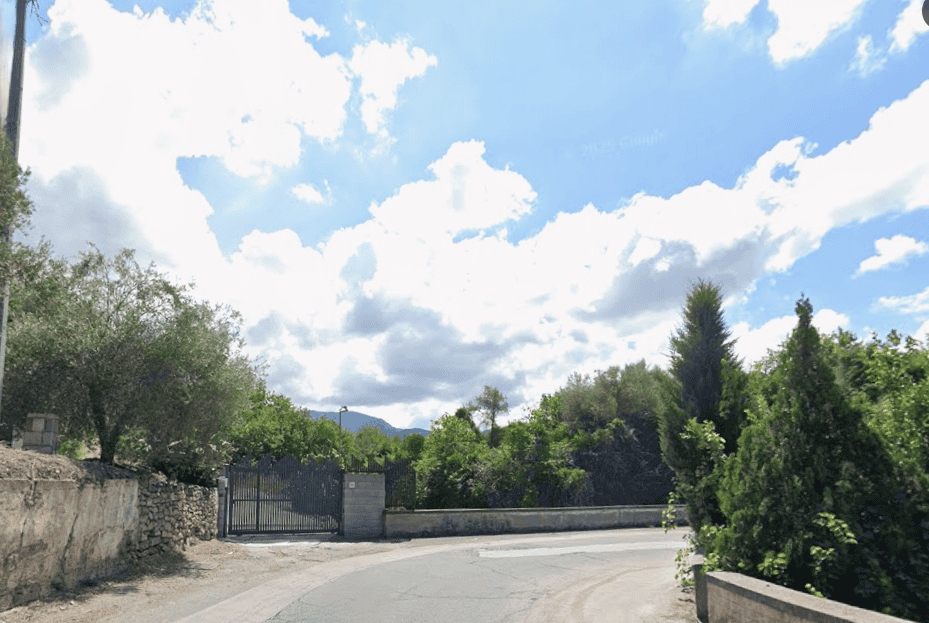 Viale di campagna con cielo sereno e nuvole bianche, alberi verdi e siepi lungo la strada, ingresso di una proprietà privata con cancello in ferro, paesaggio naturale e tranquillo.