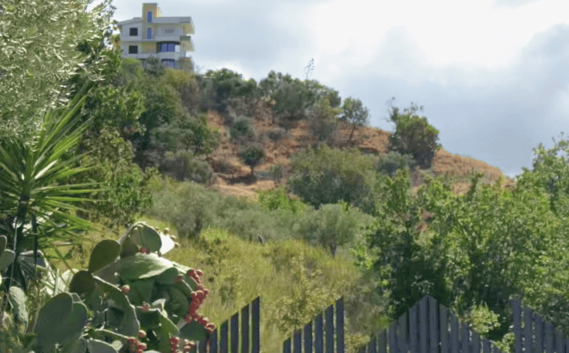 Vista panoramica di una collina con vegetazione mediterranea e una moderna casa bianca in alto, in un paesaggio naturale e tranquillo.