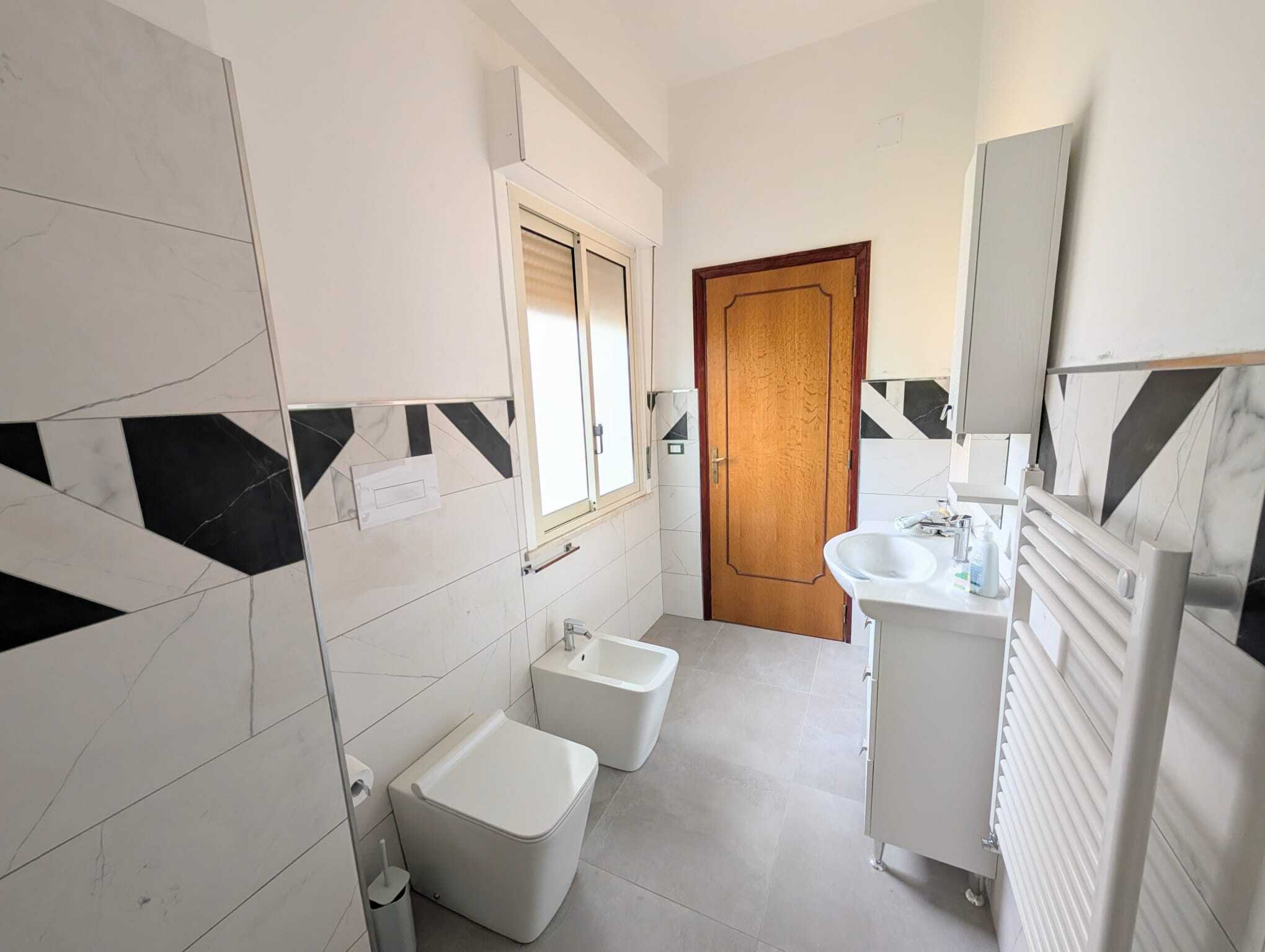 Lavabo e bidet moderni in un bagno luminoso con pareti bianche e dettagli in nero, finestra con persiana, porta in legno e radiatore asciugamani.