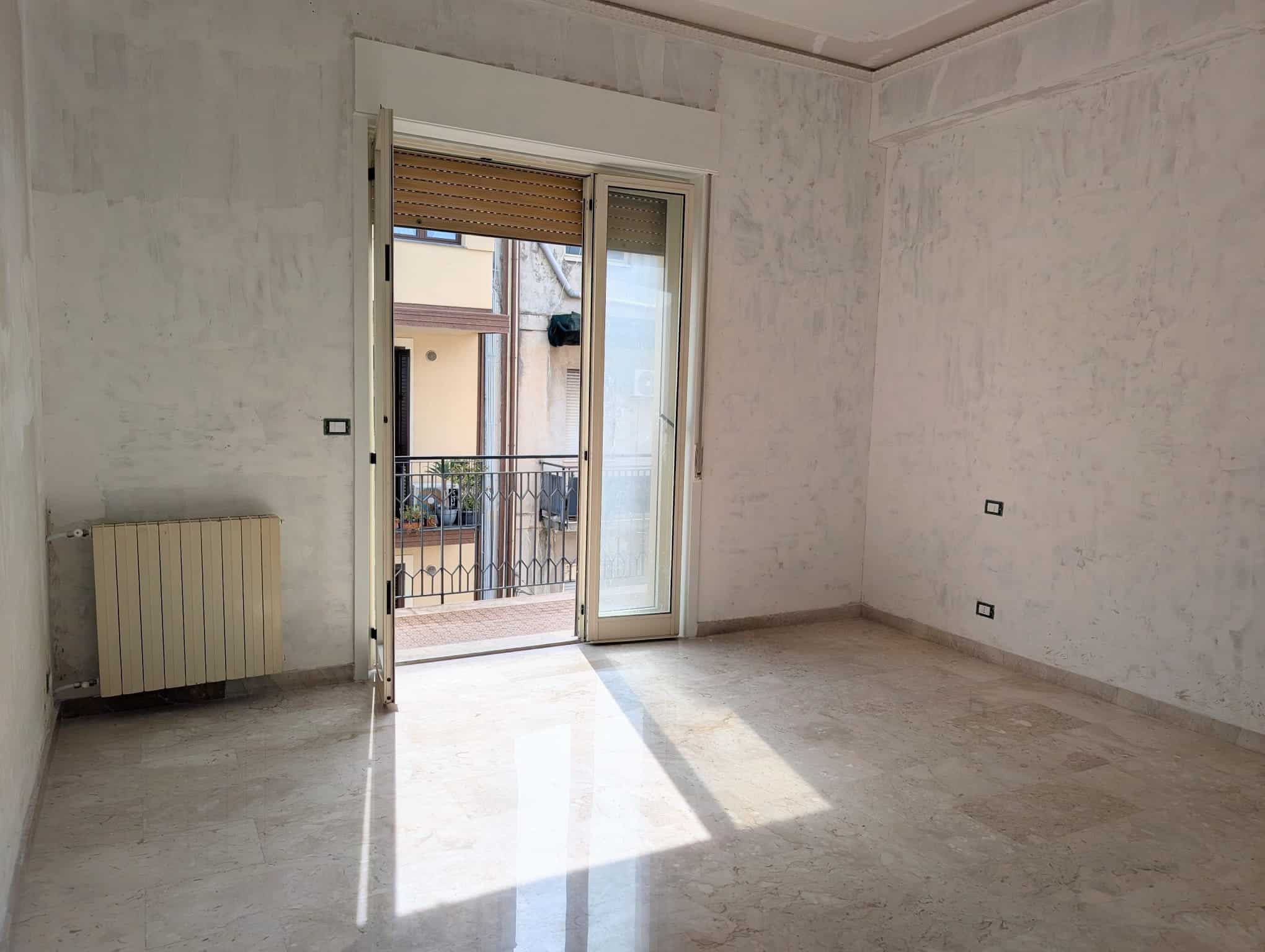 Stanza vuota in fase di ristrutturazione con porta finestra che apre su un balcone, pareti e pavimenti in fase di finitura, ambiente luminoso e spazioso.