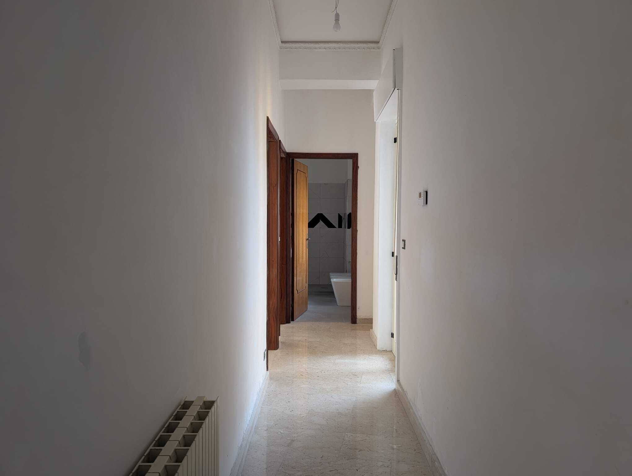 Corridoio interno di un appartamento con pareti bianche, pavimento in marmo chiaro e porta in legno, vista verso il bagno con sanitari visibili sullo sfondo.