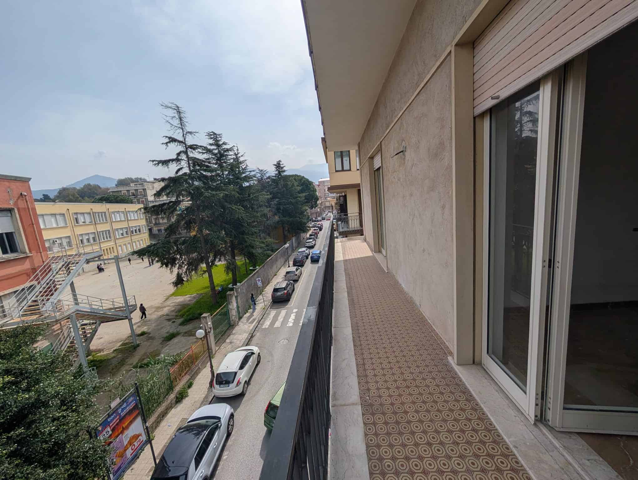 Balcone di un appartamento con vista sulla strada e sugli edifici circostanti, con panchina e piante verdi, in un contesto urbano residenziale.