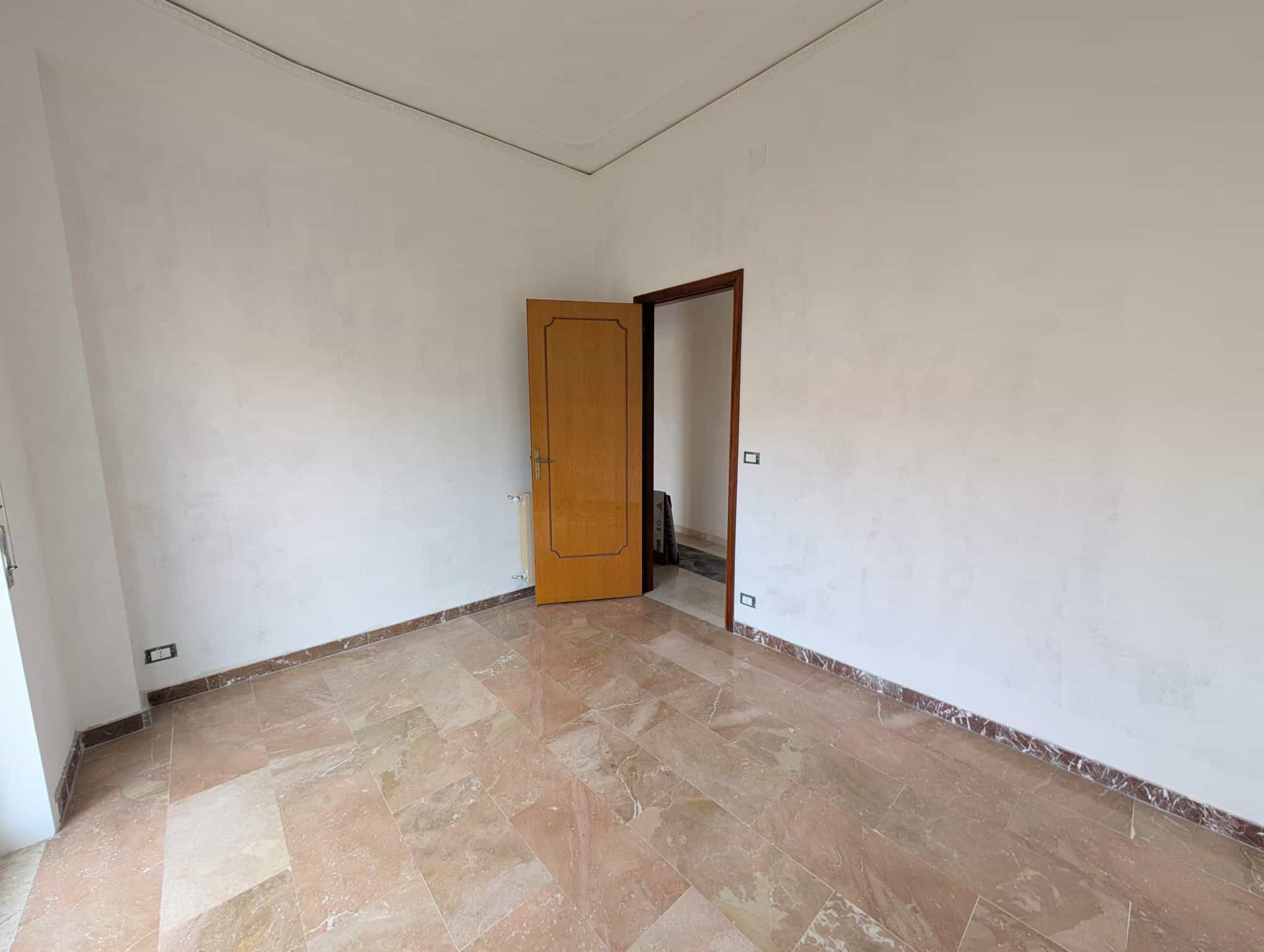 Ampia stanza vuota con pareti bianche e pavimento in marmo beige, porta in legno aperta che conduce a un'altra stanza, ideale per ristrutturazione e personalizzazione abitativa.