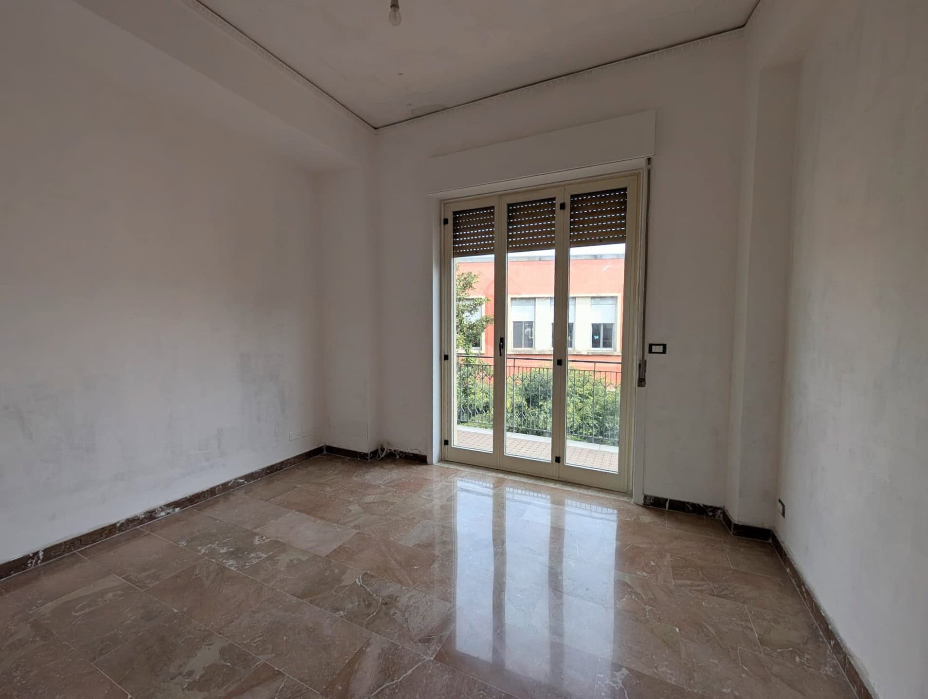 Stanza vuota in fase di ristrutturazione con grande finestra e vista su balcone e giardino, pronta per arredare e personalizzare.