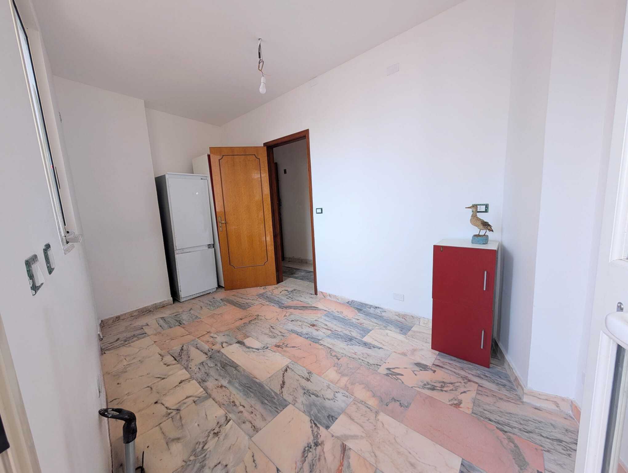 Ampio spazio vuoto in cucina con pavimento in marmo, pareti bianche e porta in legno, frigorifero bianco e mobile rosso con lampada da tavolo, luce naturale e ambiente luminoso.