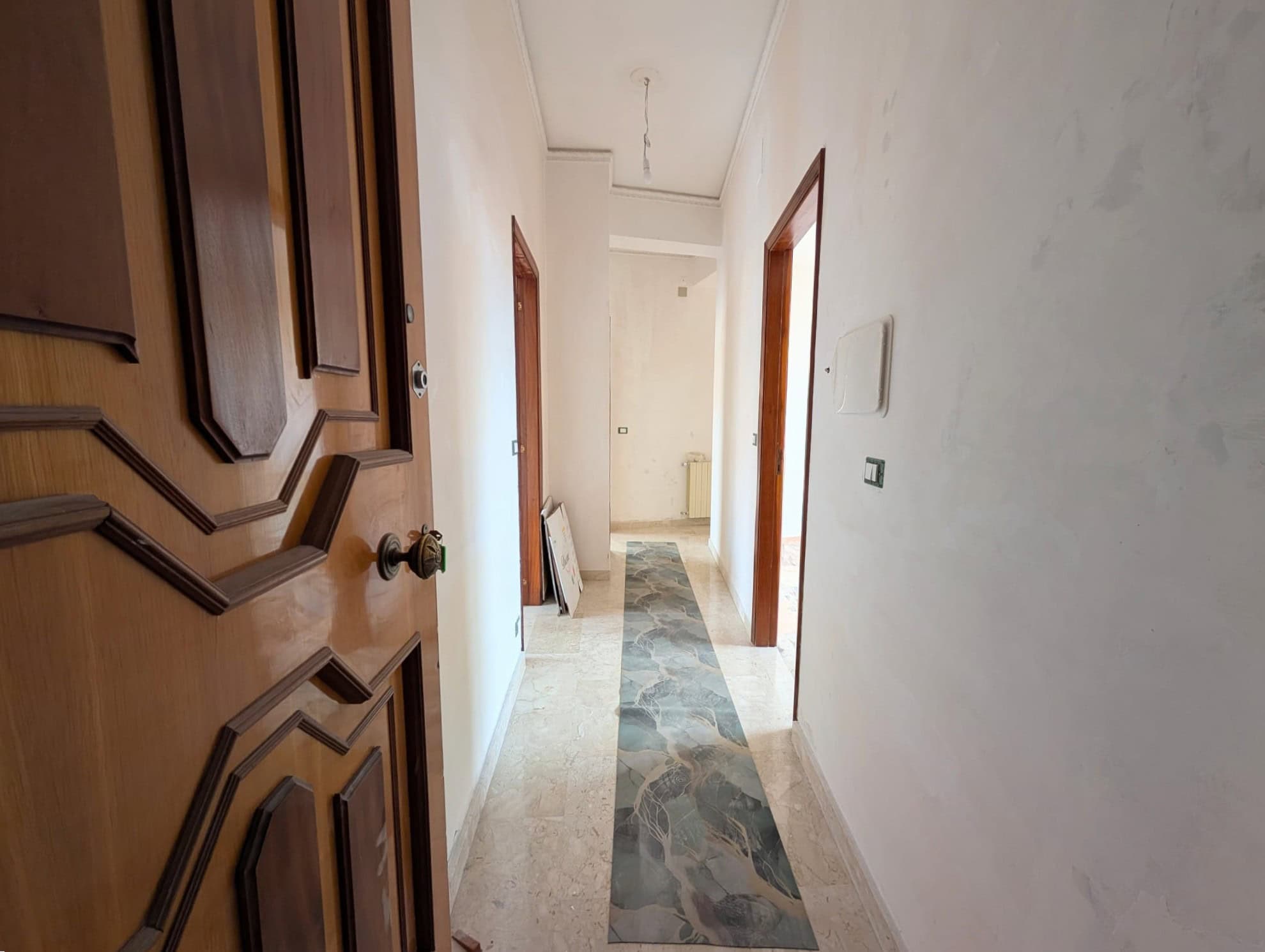 Corridoio interno di un appartamento con pareti bianche, porta in legno e pavimento in marmo, luce naturale e spazi vuoti, ideale per immobili residenziali in vendita o affitto.