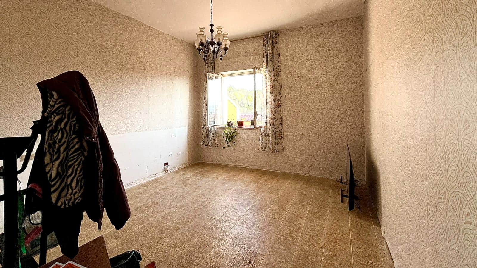 Spazio vuoto in una stanza luminosa con pareti decorate, grande finestra con tende floreali e lampadario centrale, pronta per essere arredata e personalizzata.