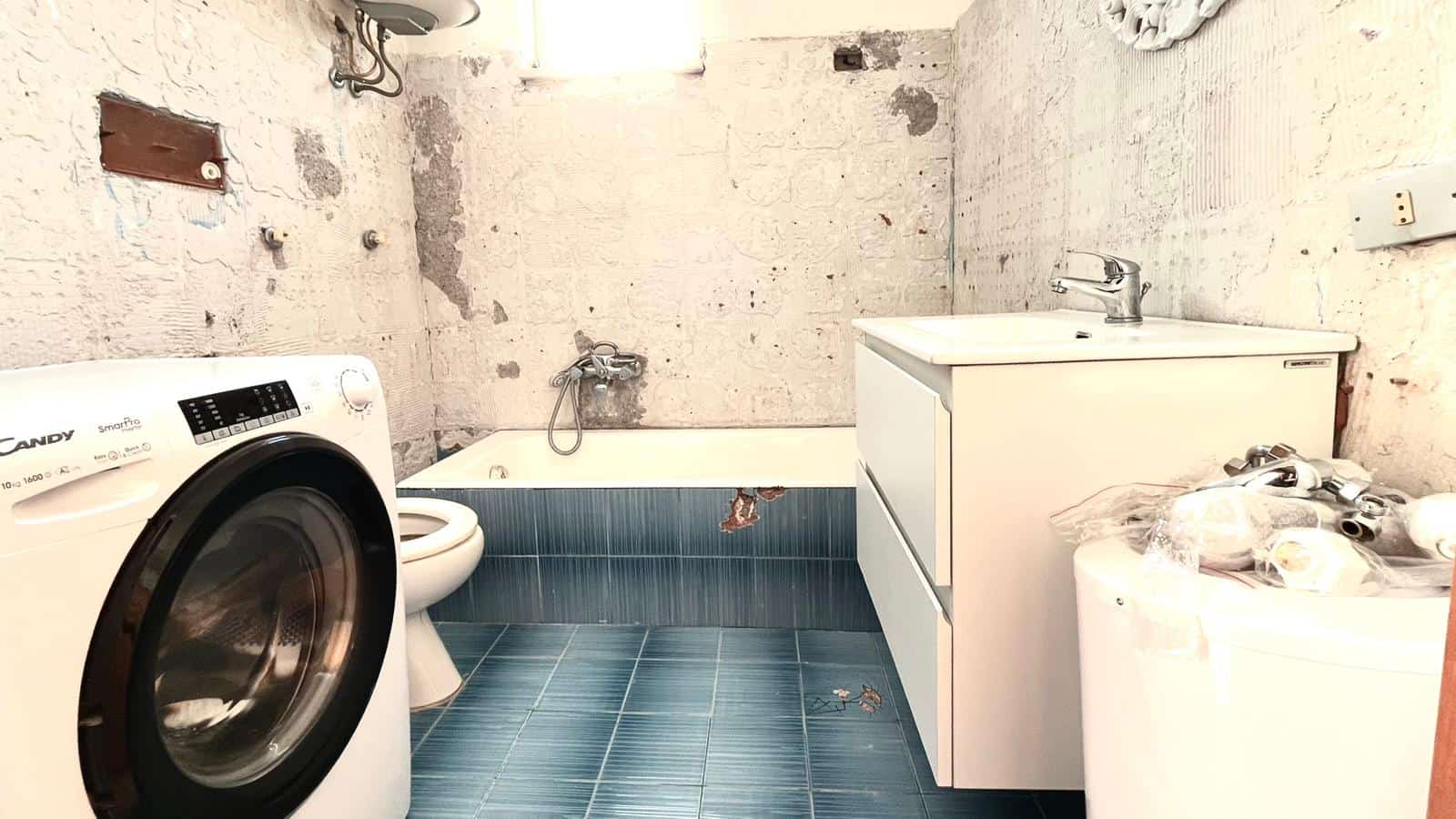 Lavatrice e bagno con vasca in un ambiente ristrutturato, pareti in cemento e pavimento in piastrelle blu, spazio funzionale e moderno per le faccende domestiche.