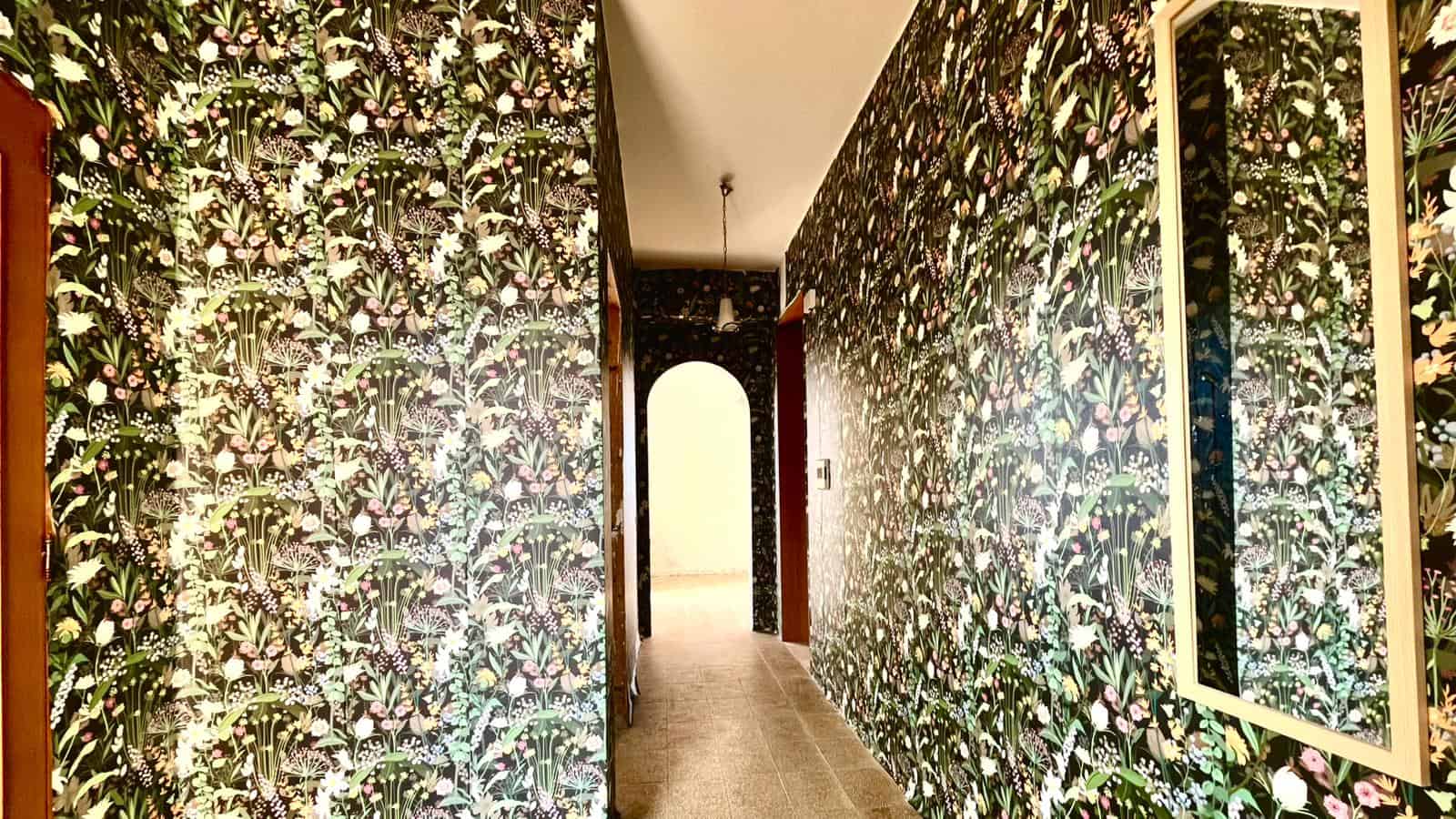 Elegante corridoio interno con pareti rivestite da carta da parati floreale, illuminato da luce naturale e lampada a soffitto, in un ambiente raffinato e accogliente.