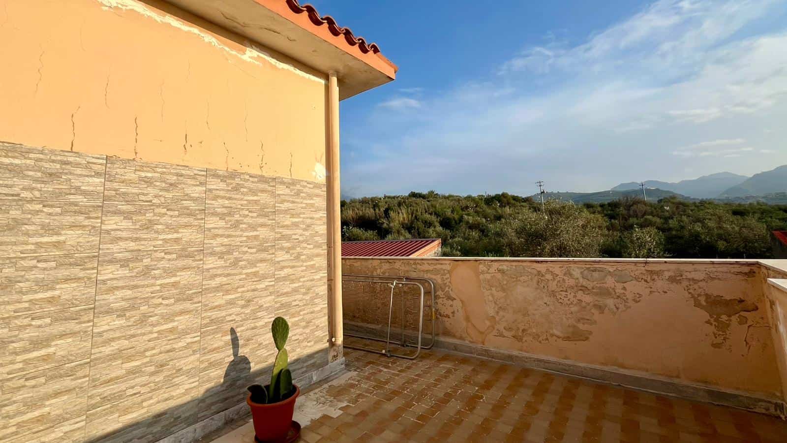 Balcone con vista panoramica sulle colline e cielo sereno, rivestimento in pietra e parete in stucco color pesca, con pianta di cactus in vaso rosso, ideale per relax e momenti all'aperto.
