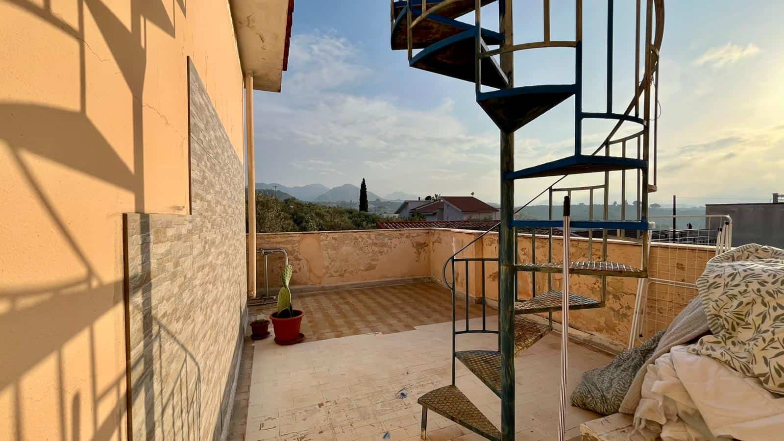Vista panoramica di un terrazzo con scala a chiocciola in metallo, arredato con piante e tessuti, con sfondo di colline e cielo sereno, ideale per relax e convivialità all'aperto.