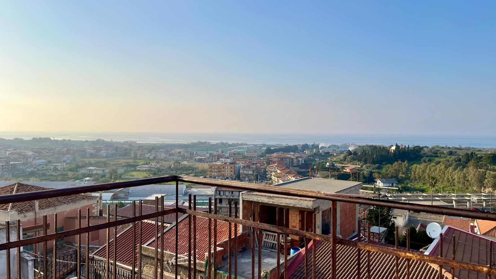 Vista panoramica sulla città e sul mare dal balcone di un appartamento in zona residenziale, con ringhiera in ferro battuto e tetto in tegole rosse, al tramonto.