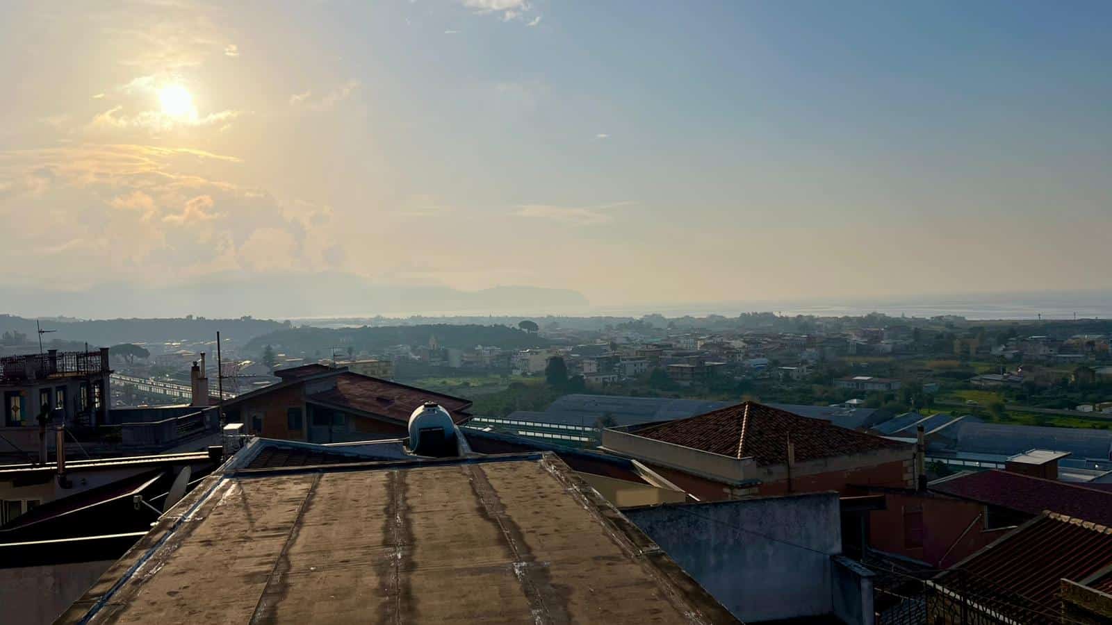 Aggiungi una vista panoramica di un quartiere residenziale con tetti e campagna circostante al tramonto, evidenziando l'urbanistica e il paesaggio rurale.