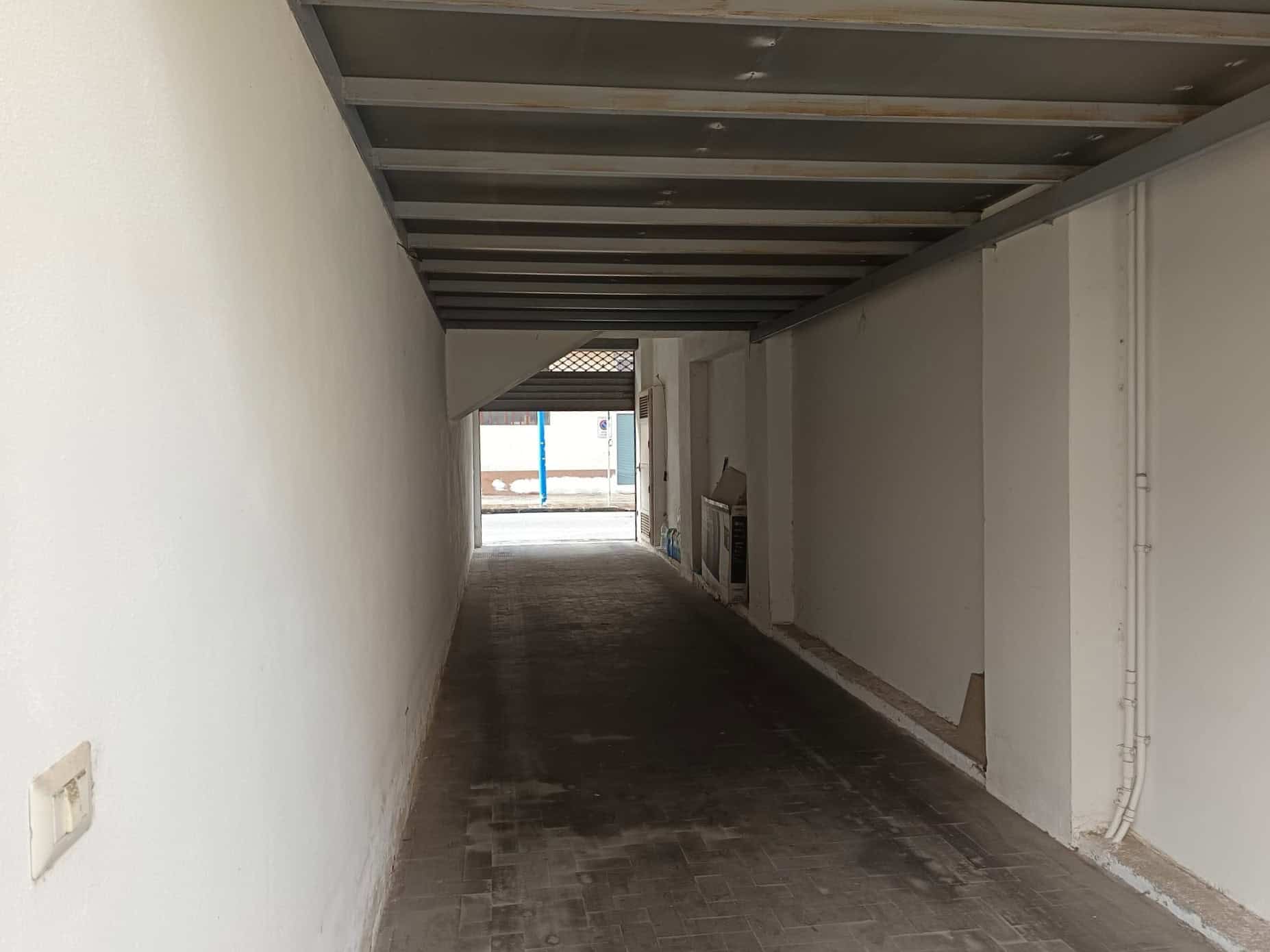Il sottopassaggio pedonale coperto con pareti bianche e pavimento in pietra, situato tra edifici, con ingresso visibile in fondo e struttura in metallo sul soffitto.