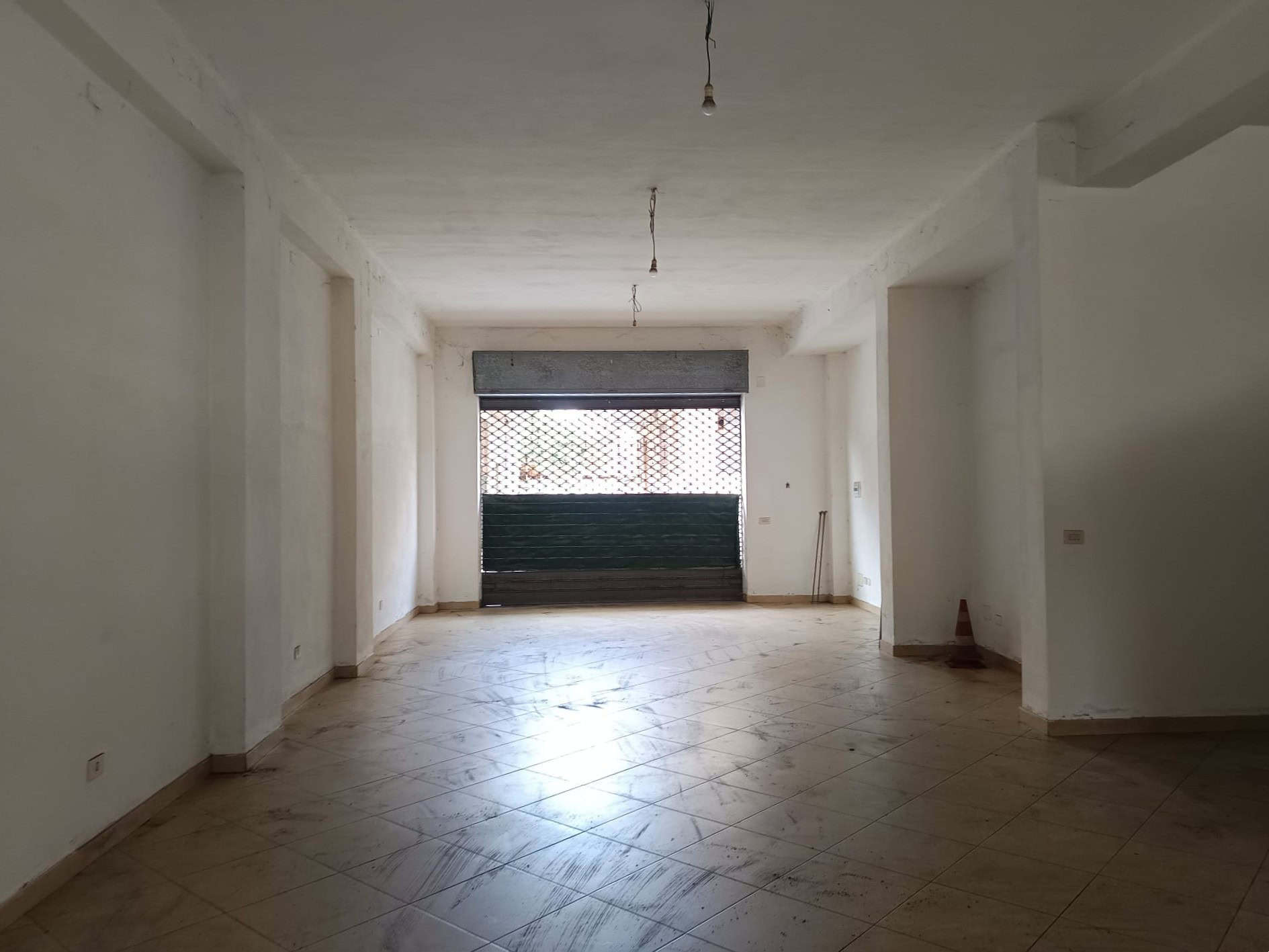 Ampio locale commerciale o ufficio vuoto con pareti bianche e grande finestra con serranda, pronto per ristrutturazione, ideale per attività o studio professionale.