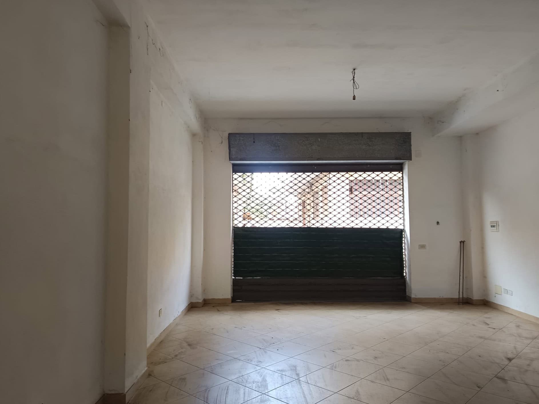 Il testo dell'immagine mostra un interno di un appartamento vuoto con pareti bianche e pavimento in piastrelle, con una grande finestra con tapparella e inferriate, pronto per essere arredato.