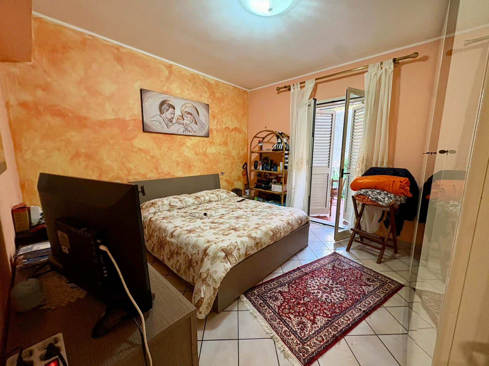 Ampio soggiorno con pareti color pesca, letto matrimoniale, televisore, balcone con tende leggere, tappeto persiano, scaffale con libri e oggetti decorativi, e una sedia con coperta arancione.