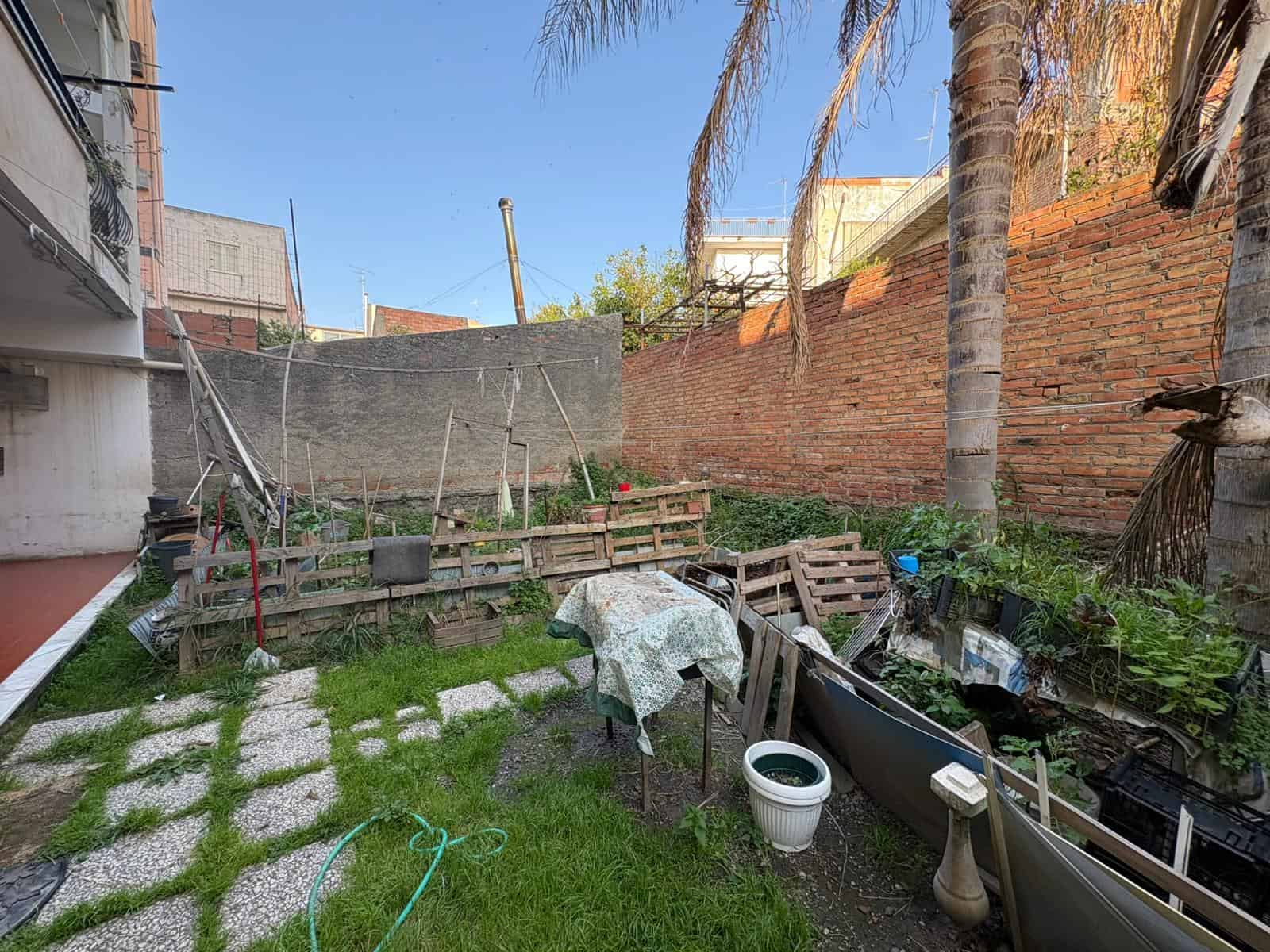 Ampio giardino urbano con piante, palme e area relax, recintato con muro di mattoni e rete protettiva, ideale per orto e svago all'aperto, in zona residenziale tranquilla.