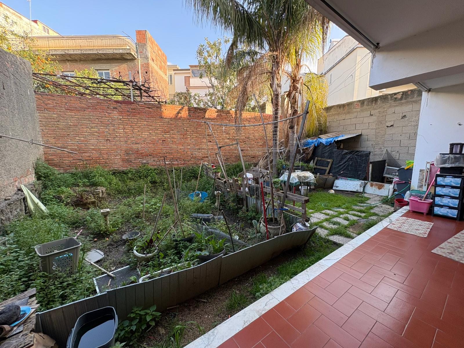 Pianta da giardino con alberi e piante in un cortile urbano, recintata con rete metallica, con area pavimentata in piastrelle rosse e spazio per coltivare ortaggi e fiori, in un ambiente residenziale.