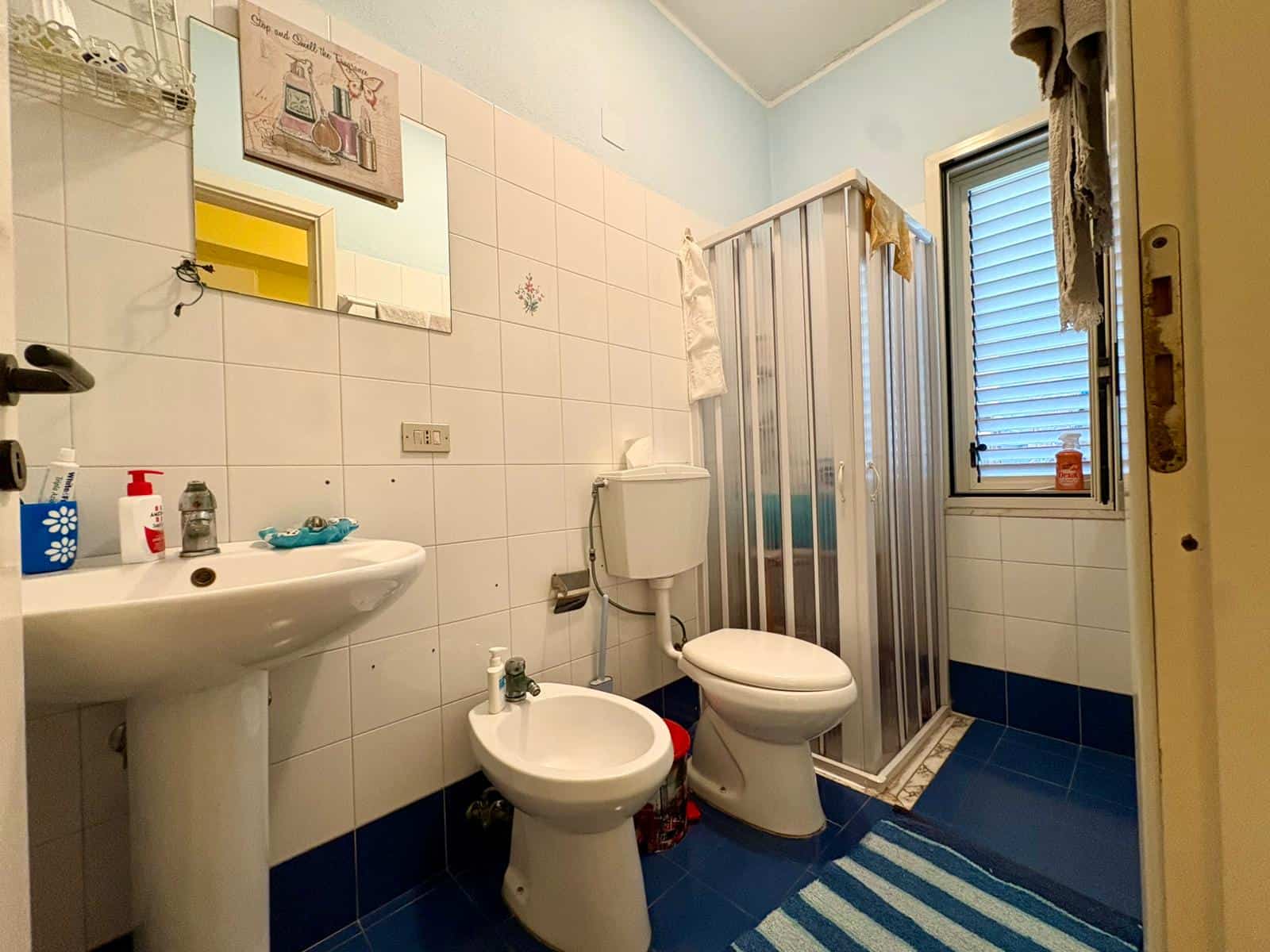 Bagno con sanitari e box doccia in un appartamento, dettagli di arredo e accessori per la pulizia, finestra con tapparelle, pavimento in piastrelle blu e pareti bianche, ambiente funzionale e compatto.