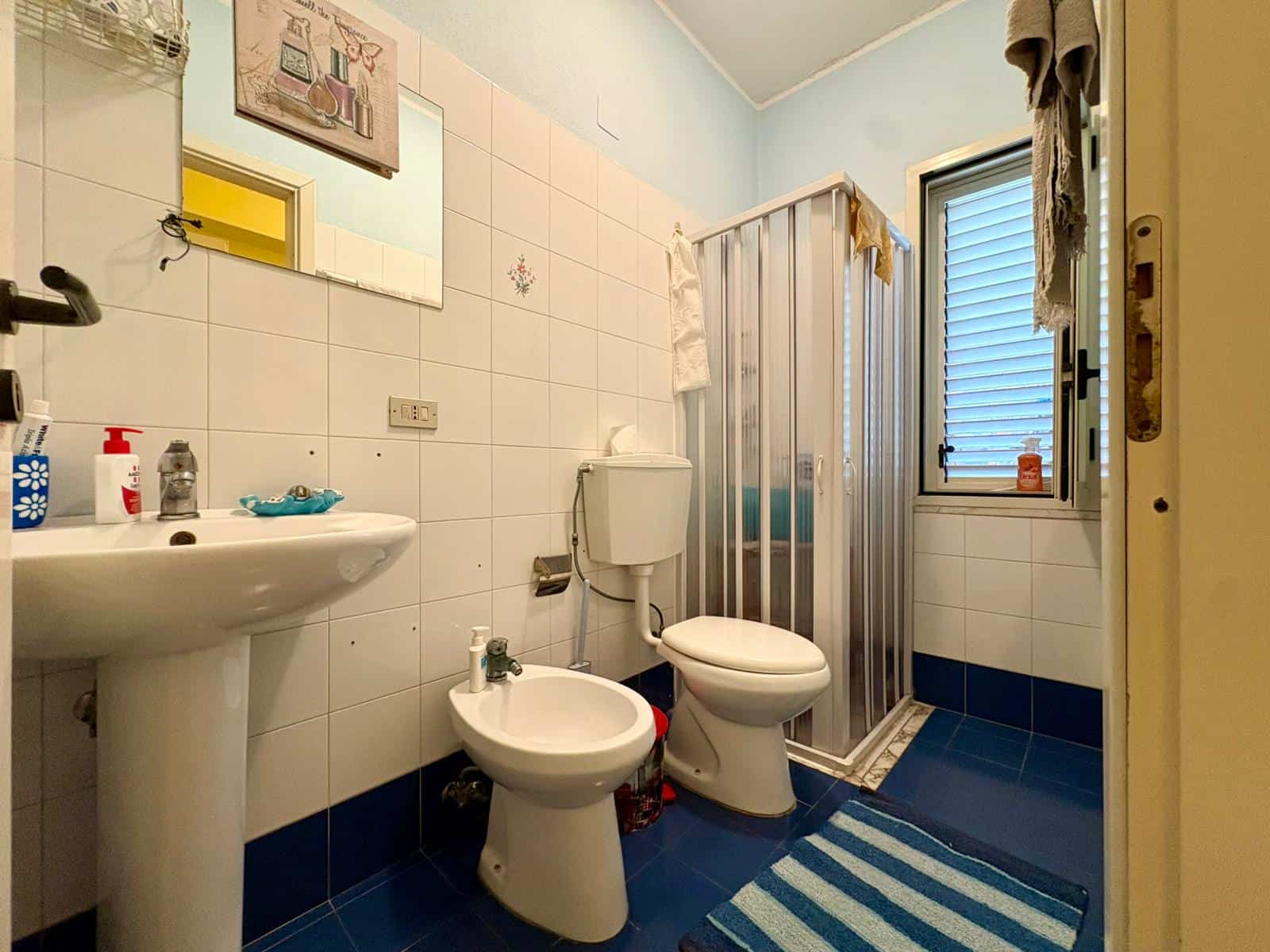 Lavabo e servizi igienici in bagno con piastrelle bianche e blu, doccia separata con porta in vetro, finestra per ventilazione naturale, accessori essenziali e tappetino antiscivolo.
