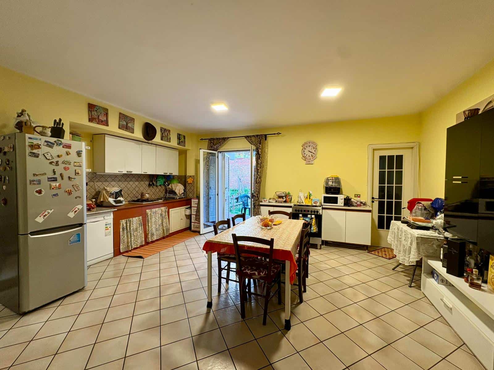 Cucina spaziosa e luminosa con tavolo centrale, frigorifero, armadietti bianchi e accessori vari, ambiente accogliente e funzionale.