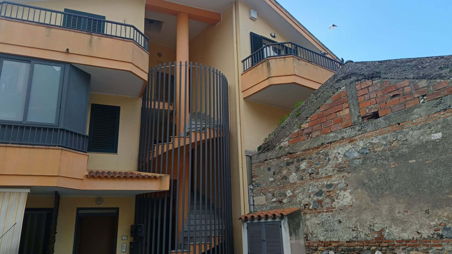 Escalatore esterno in metallo nero che collega i piani di un edificio residenziale con balconi e finestre, in un contesto urbano con pareti in mattoni e facciata color beige.
