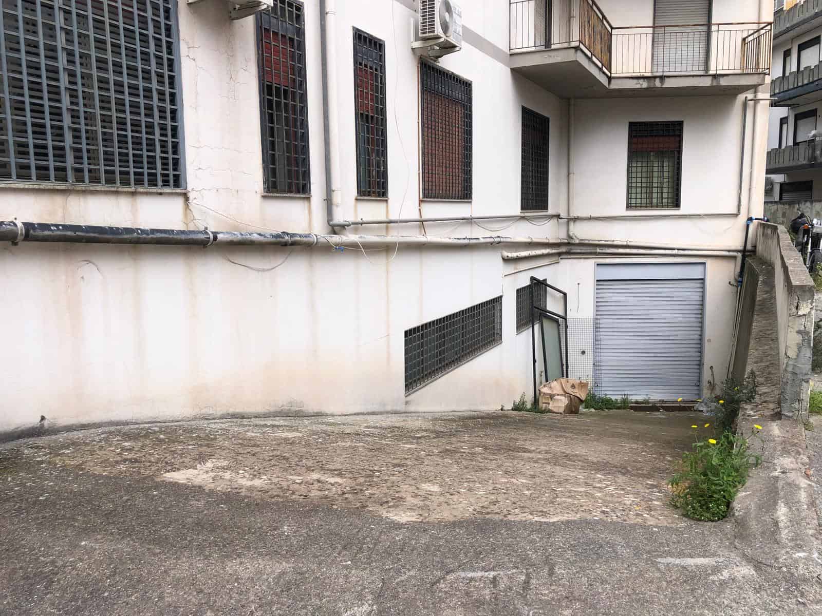 Vano cortile condominiale con pavimentazione in cemento e pareti bianche, accesso a garage con serranda metallica, situato in un contesto urbano residenziale a NextCasa.