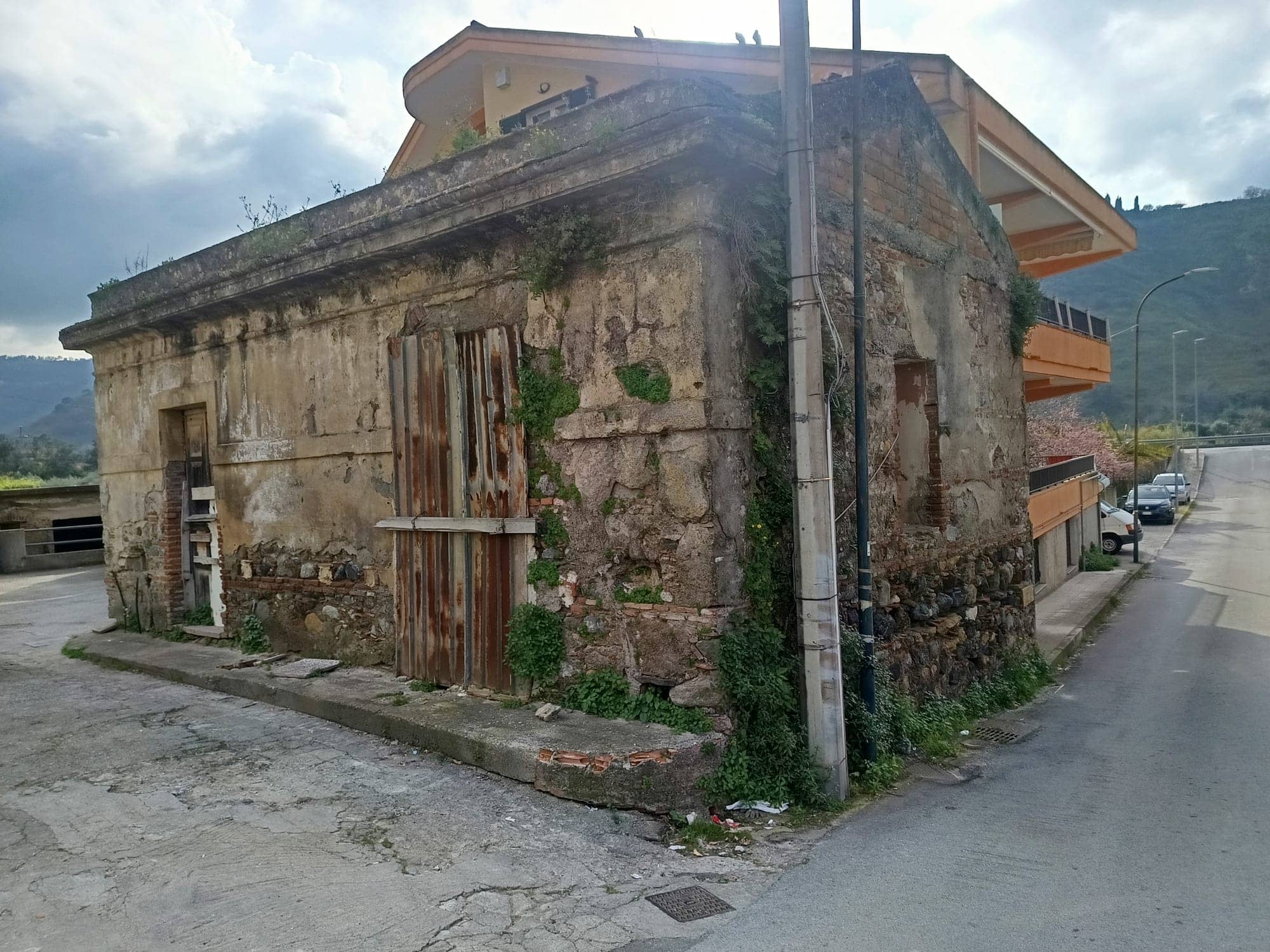 Vecchio edificio in pietra e mattoni con porte e finestre in legno, situato in un'area urbana con montagne sullo sfondo, rappresenta un esempio di architettura storica e degrado urbano.