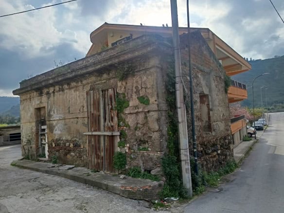 Vecchio edificio in pietra con porte di legno, situato in un'area urbana vicino a moderni appartamenti e strade asfaltate, con montagne sullo sfondo e cielo nuvoloso.