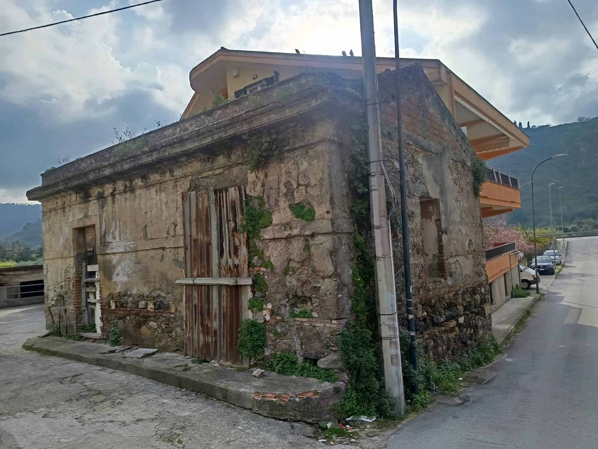 Vecchio edificio in pietra con porte di legno, situato in un'area urbana vicino a moderni appartamenti e strade asfaltate, con montagne sullo sfondo e cielo nuvoloso.