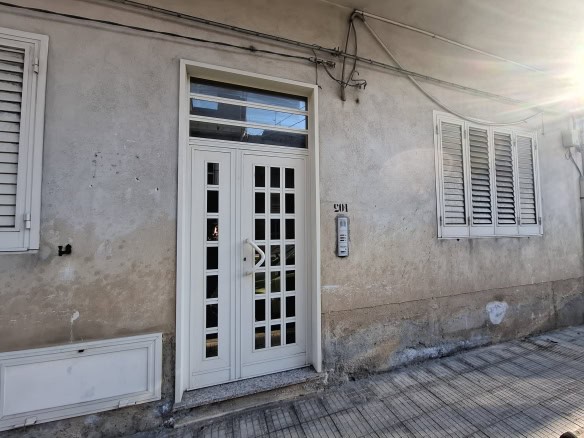 Porta d'ingresso bianca con vetri decorativi e finestra con persiane in legno, facciata esterna di una casa in stile semplice, illuminata dal sole, in un contesto urbano.
