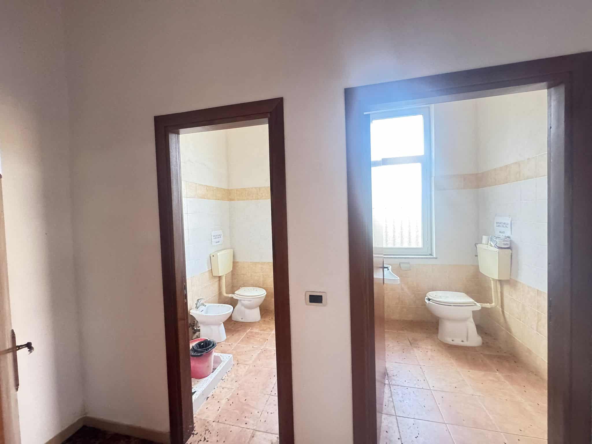 Bagni pubblici con pavimenti in piastrelle e sanitari moderni, illuminati dalla luce naturale proveniente da una grande finestra, ideali per strutture pubbliche o commerciali.
