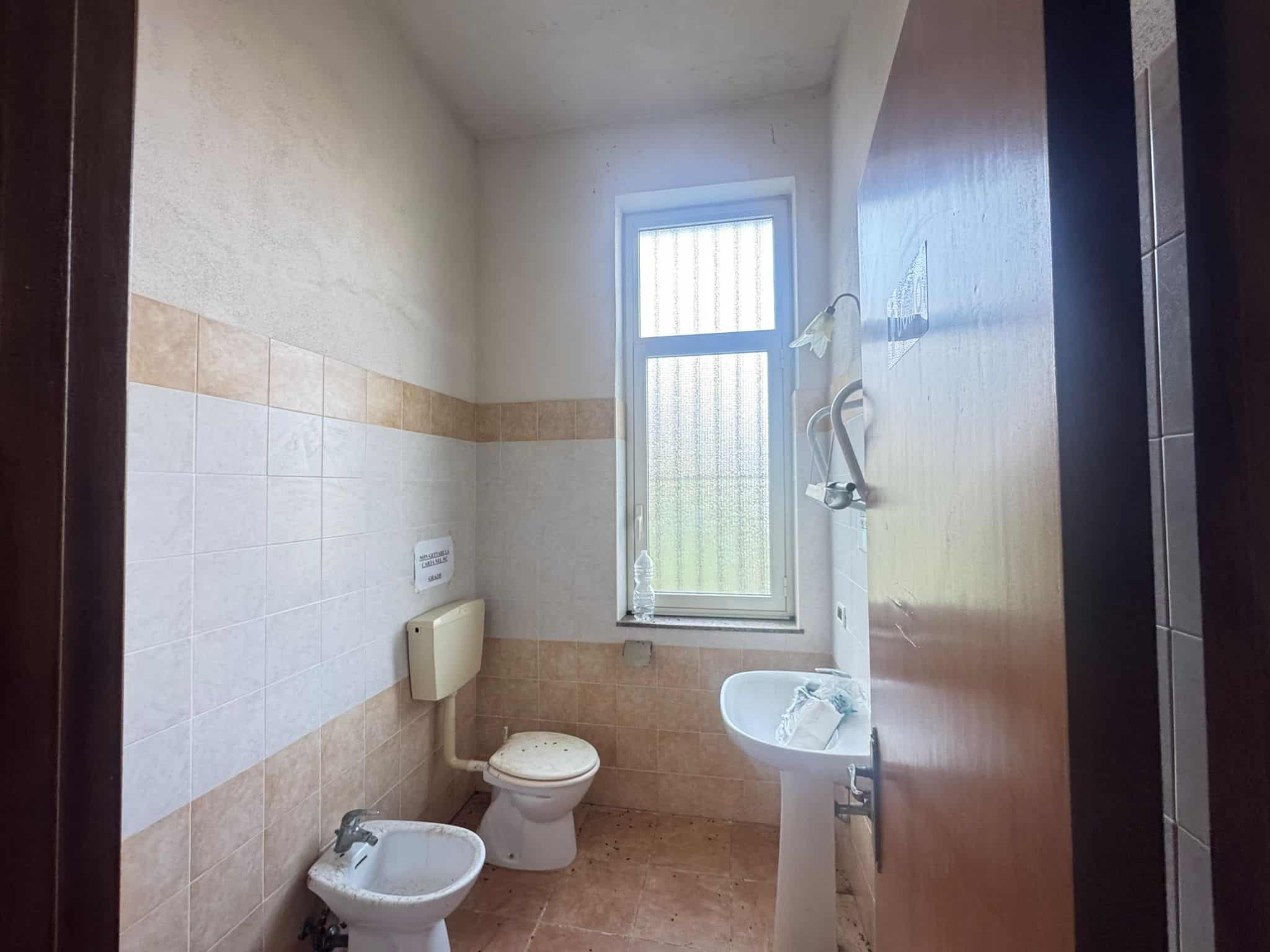 Lavabo e servizi igienici in un bagno di piccole dimensioni con finestra, piastrelle beige e pareti bianche, ideale per appartamenti o case compatte.